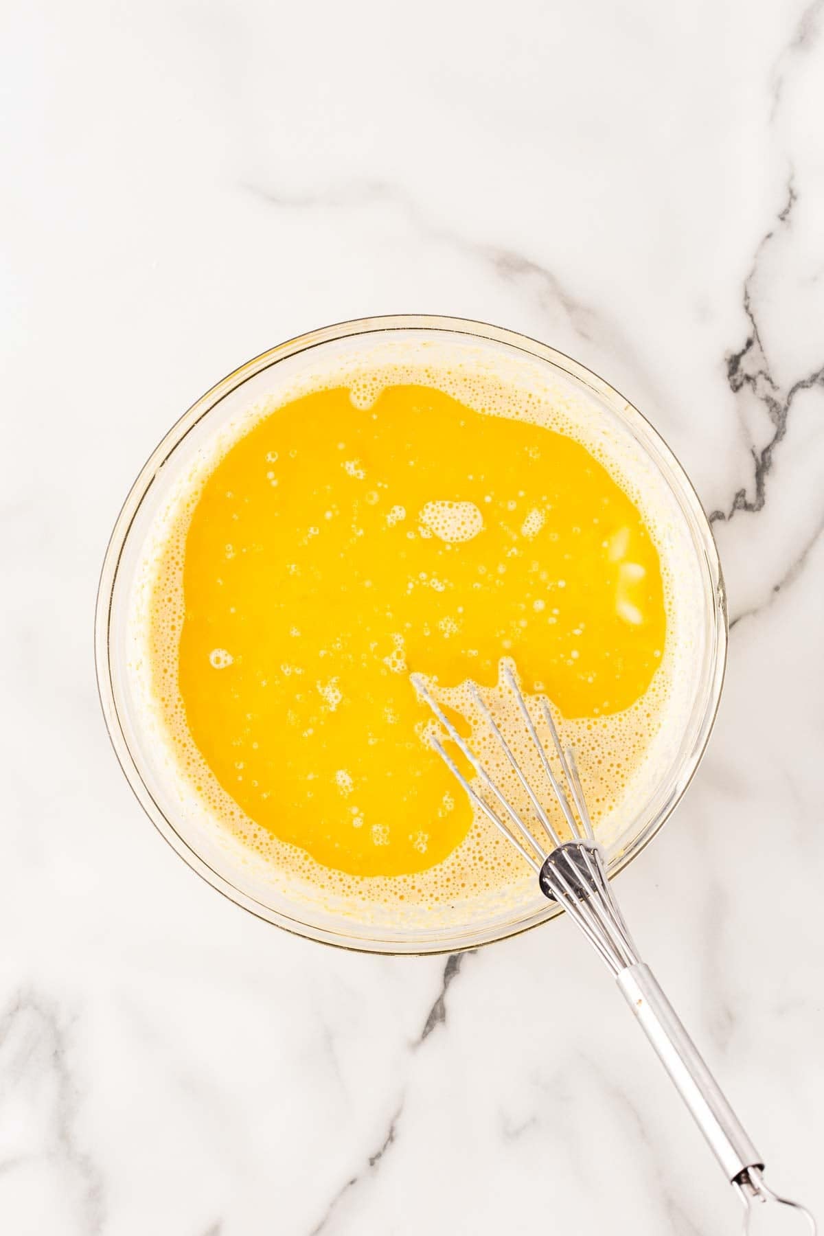 Yellow cornbread batter being mixed with a metal whisk in a clear glass bowl on a marble countertop