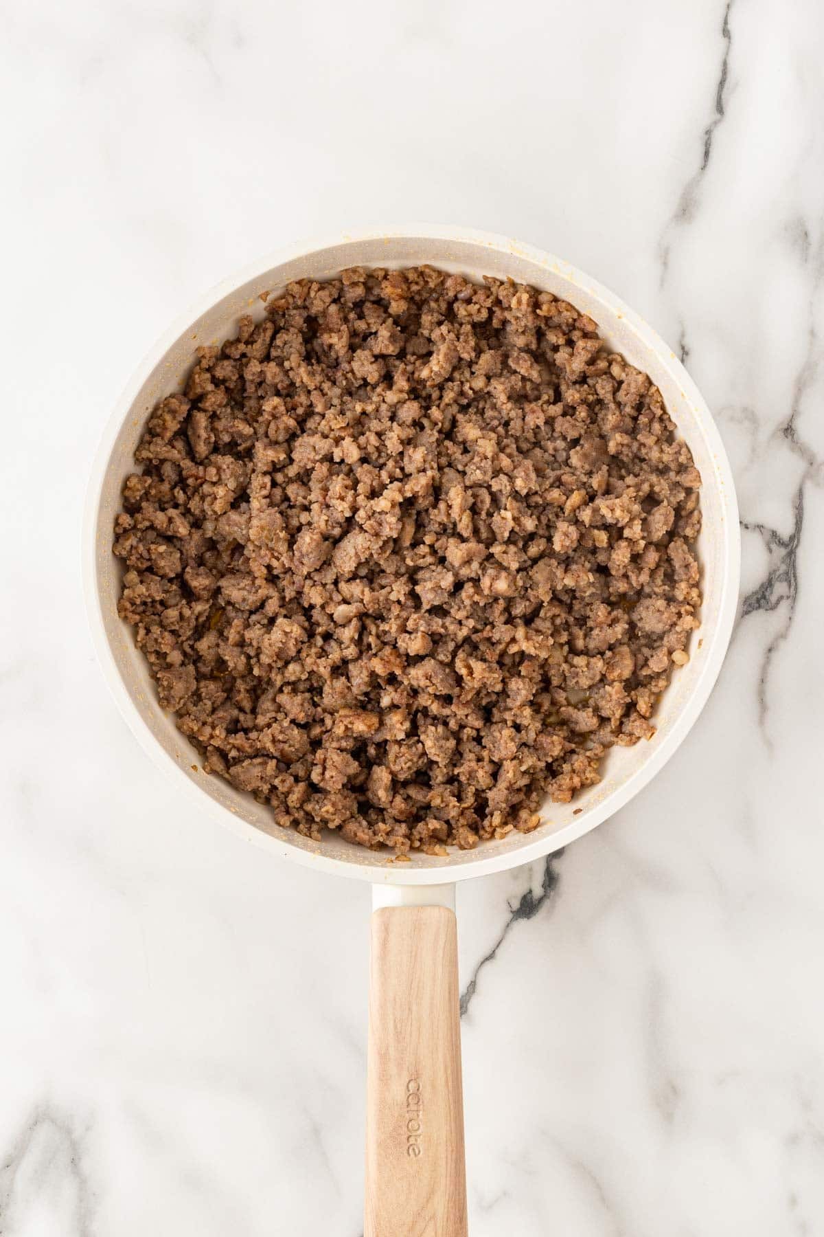 Browned sage sausage crumbles in a white skillet with a wooden handle on a marble countertop