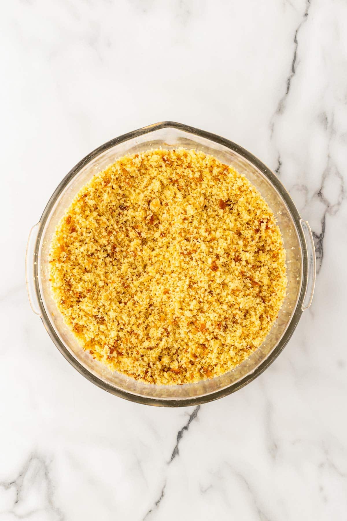 Finely crumbled golden cornbread in a large clear glass mixing bowl on a marble countertop