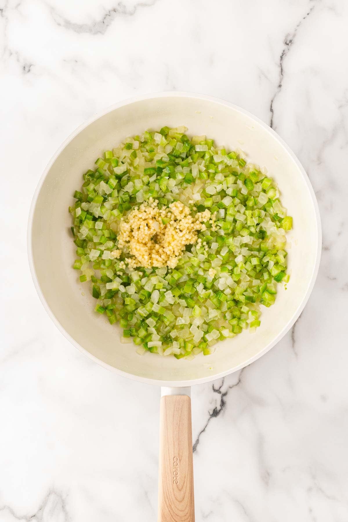 Sautéed onion, celery, green bell pepper, and minced garlic in a white skillet on a marble countertop
