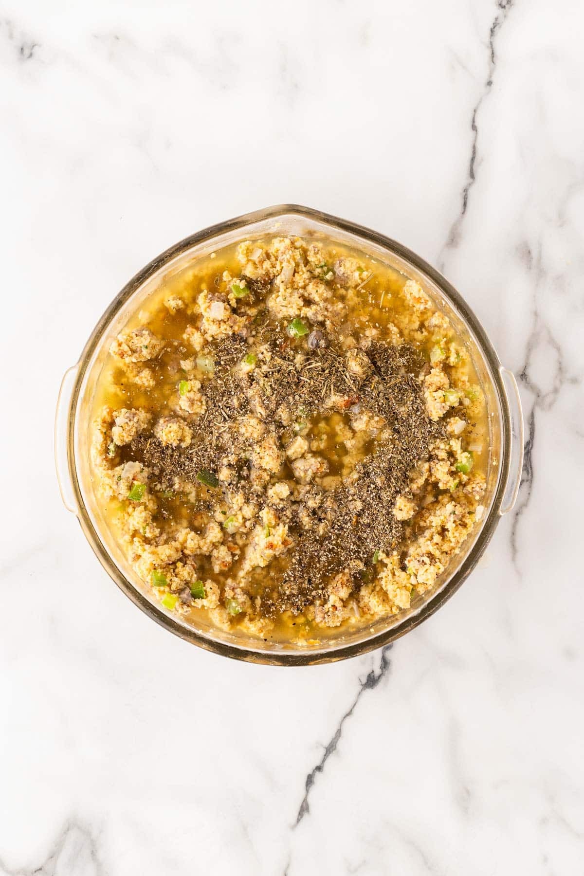 Cornbread dressing mixture topped with poultry seasoning, herbs, black pepper, and chicken broth in a glass bowl