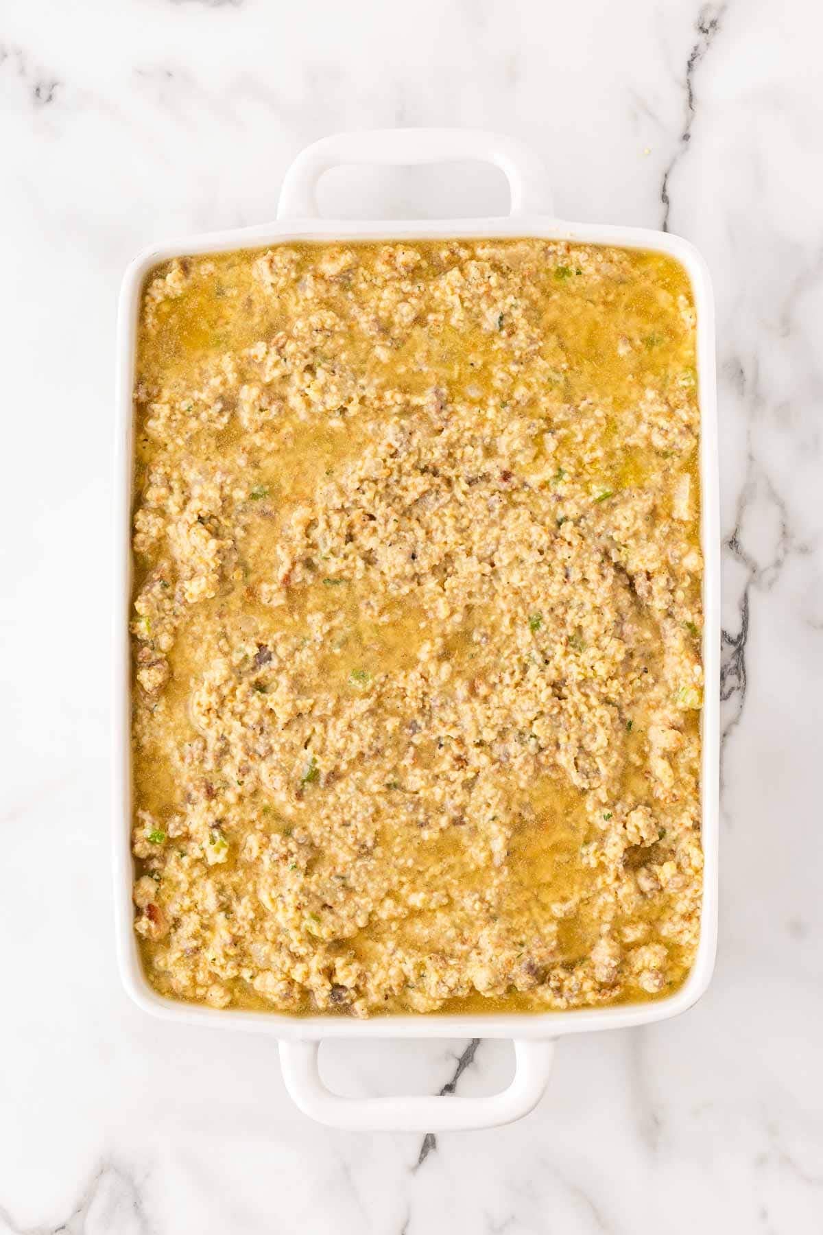 Unbaked sausage cornbread dressing with visible chicken broth in a white rectangular baking dish on a marble countertop