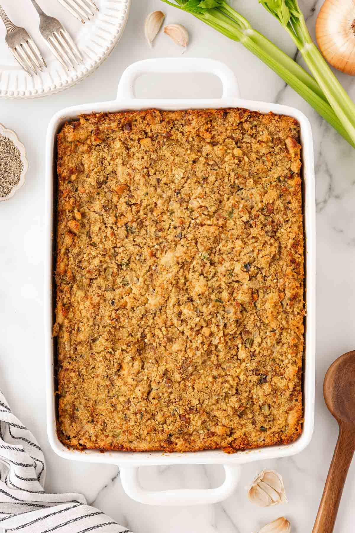 Golden brown sausage cornbread dressing in a white baking dish, surrounded by celery, onion, garlic, and silver forks on a marble countertop