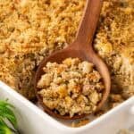 Wooden spoon scooping sausage cornbread dressing from a white baking dish, showing the moist interior and golden crust, with celery and garlic nearby