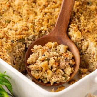 Wooden spoon scooping sausage cornbread dressing from a white baking dish, showing the moist interior and golden crust, with celery and garlic nearby