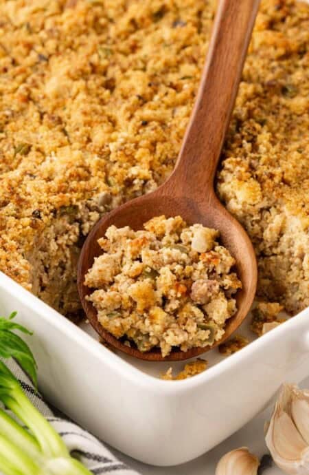 Wooden spoon scooping sausage cornbread dressing from a white baking dish, showing the moist interior and golden crust, with celery and garlic nearby