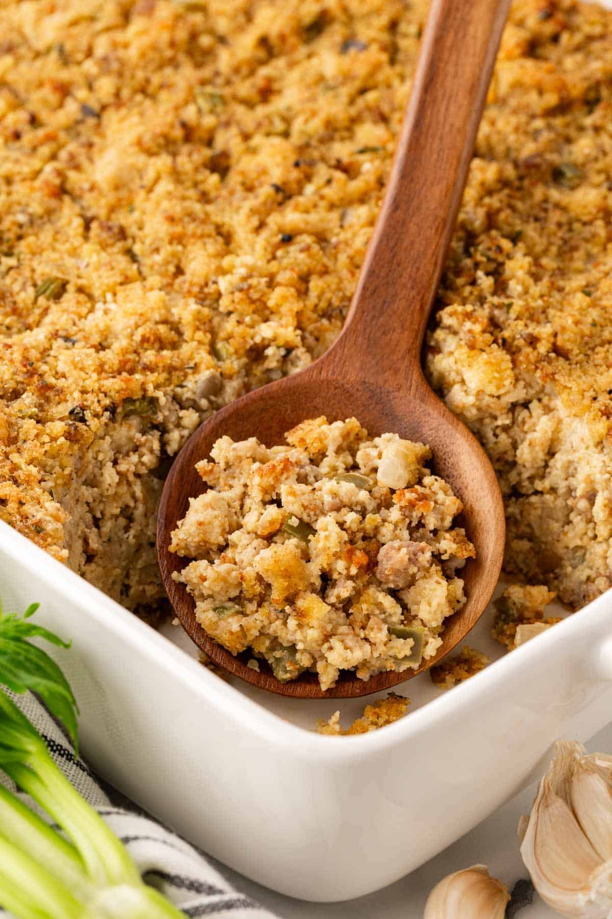 Wooden spoon scooping sausage cornbread dressing from a white baking dish, showing the moist interior and golden crust, with celery and garlic nearby