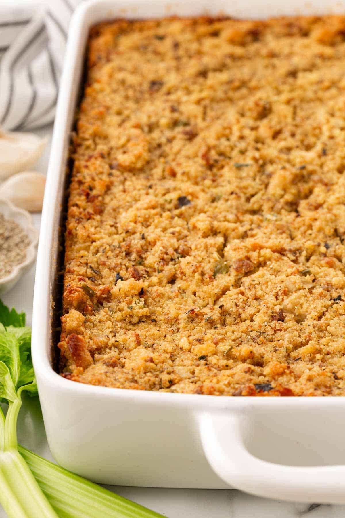 Closeup of golden brown sausage cornbread dressing in a white baking dish, with fresh celery and a striped kitchen towel nearby